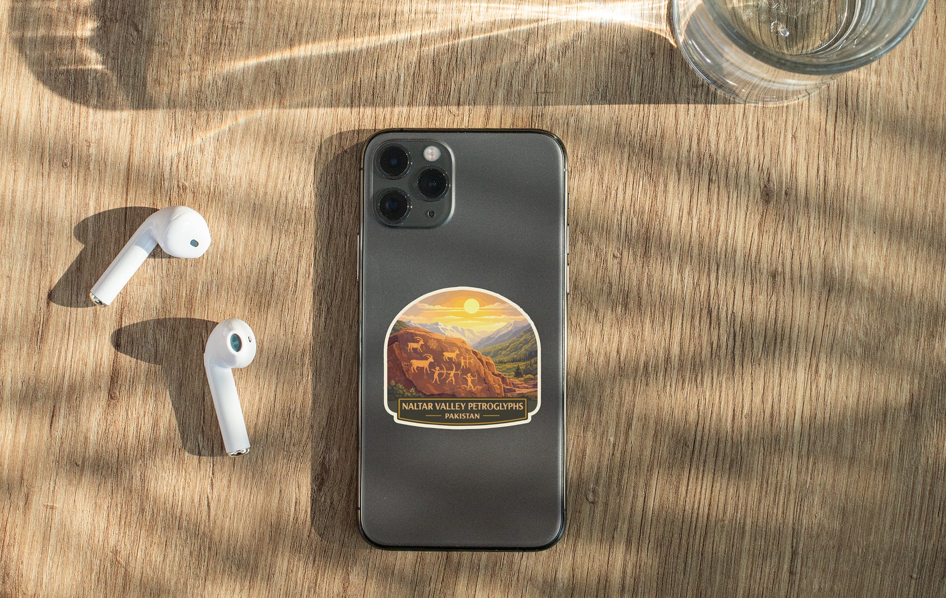 a smartphone with a sticker on the back, a pair of white earbuds, and a glass of water on a wooden surface.