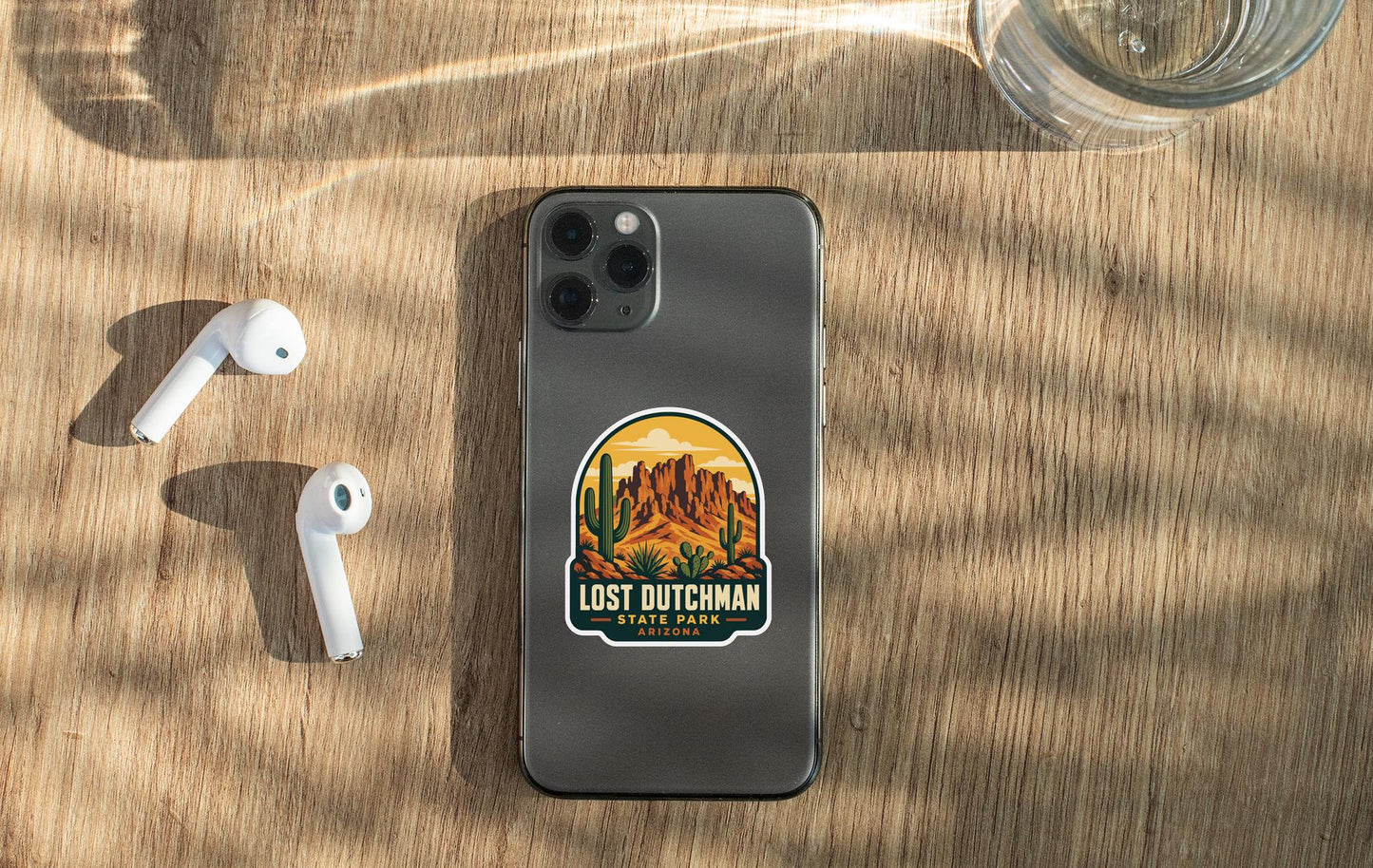 a smartphone with a sticker on it, placed on a wooden surface alongside a pair of white earbuds and a glass of water.