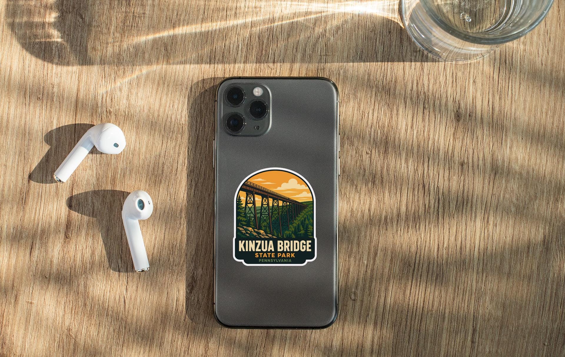 a smartphone with a sticker on the back, placed on a wooden surface alongside a pair of white earbuds and a glass of water.