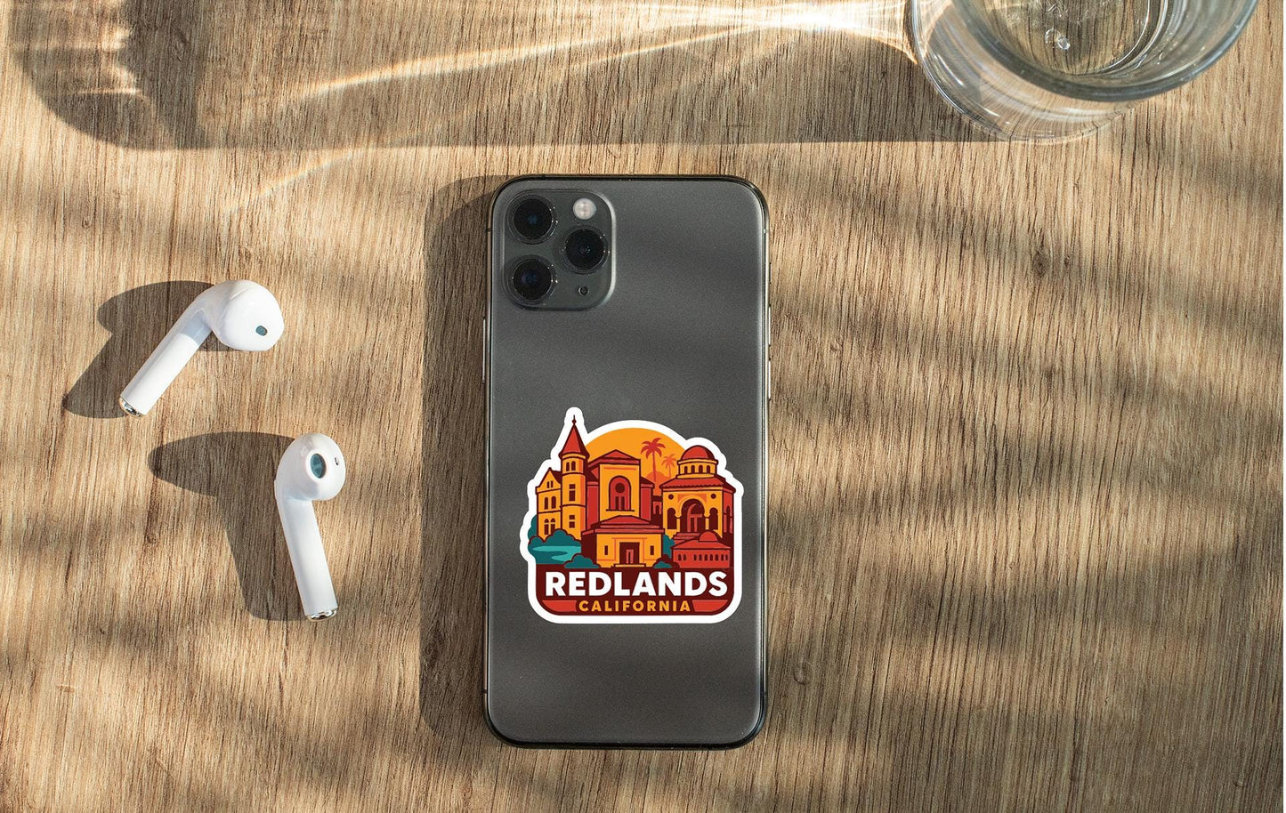 a smartphone with a sticker of a city on it, placed on a wooden surface alongside a pair of white earbuds and a glass of water.