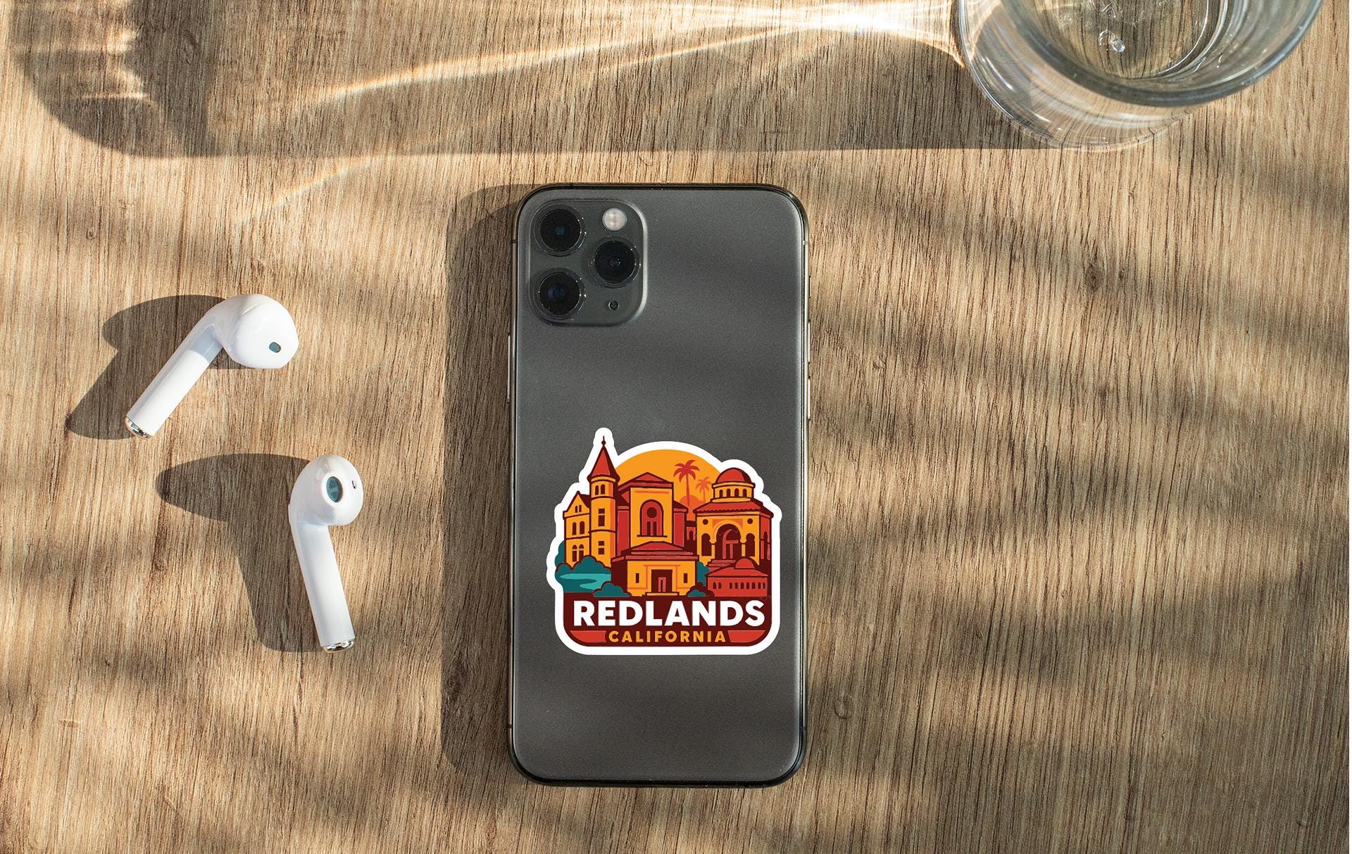 a smartphone with a sticker of a city on it, placed on a wooden surface alongside a pair of white earbuds and a glass of water.
