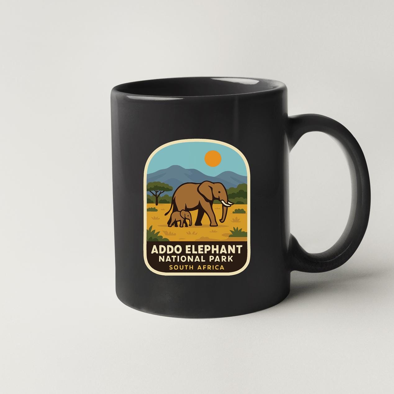 A black coffee mug with a label that features an illustration of an elephant and the text "ADDO ELEPHANT NATIONAL PARK SOUTH AFRICA".