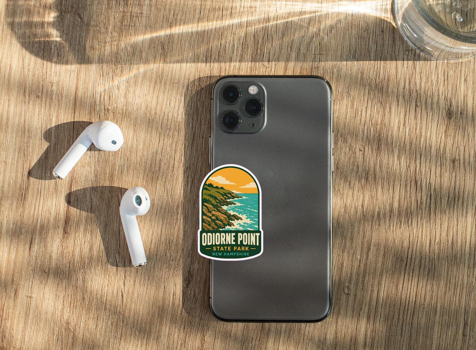 a smartphone with a sticker on it, along with a pair of white earbuds and a glass of water on a wooden surface.