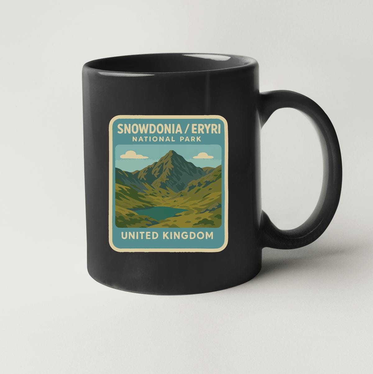 A black coffee mug with a graphic design depicting a mountain landscape and the text "SNOWDONIA/ERYRI NATIONAL PARK UNITED KINGDOM".