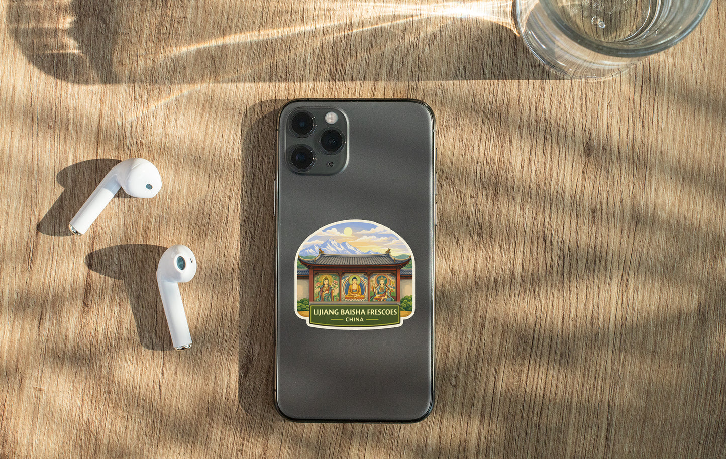 a smartphone with a sticker on it, placed on a wooden surface alongside a pair of white earbuds and a glass of water.