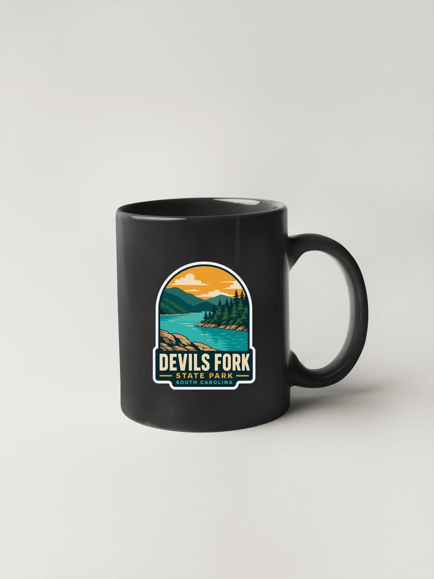 A black coffee mug with a graphic design depicting a scenic landscape, including mountains, a lake, and a forest. The text on the mug reads "DEVILS FORK STATE PARK SOUTH CAROLINA".