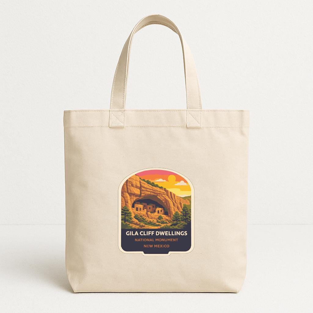 A beige tote bag with a graphic design depicting a scenic landscape, including a cave and mountains, along with text that reads "Gila Cliff Dwellings".