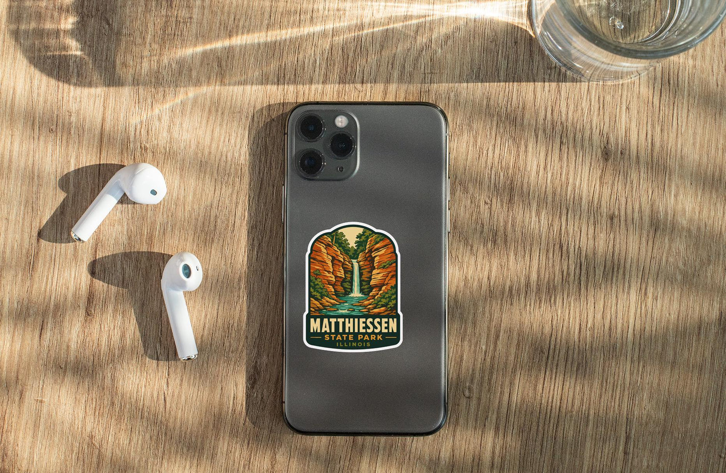 a smartphone with a sticker on the back, placed on a wooden surface alongside a pair of white earbuds and a glass of water.