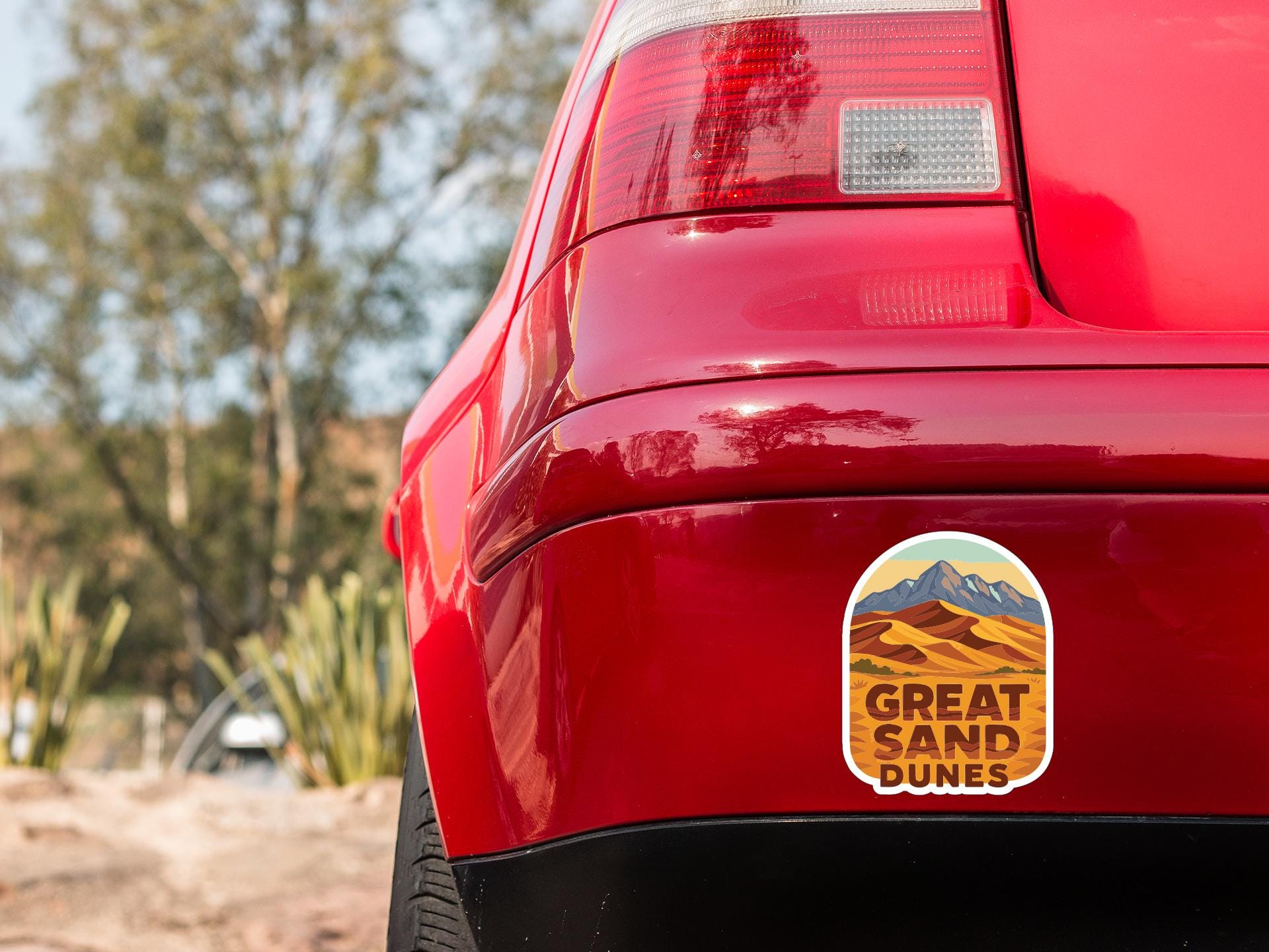 the rear end of a red car with a sticker on the bumper that says "GREAT SAND DUNES".