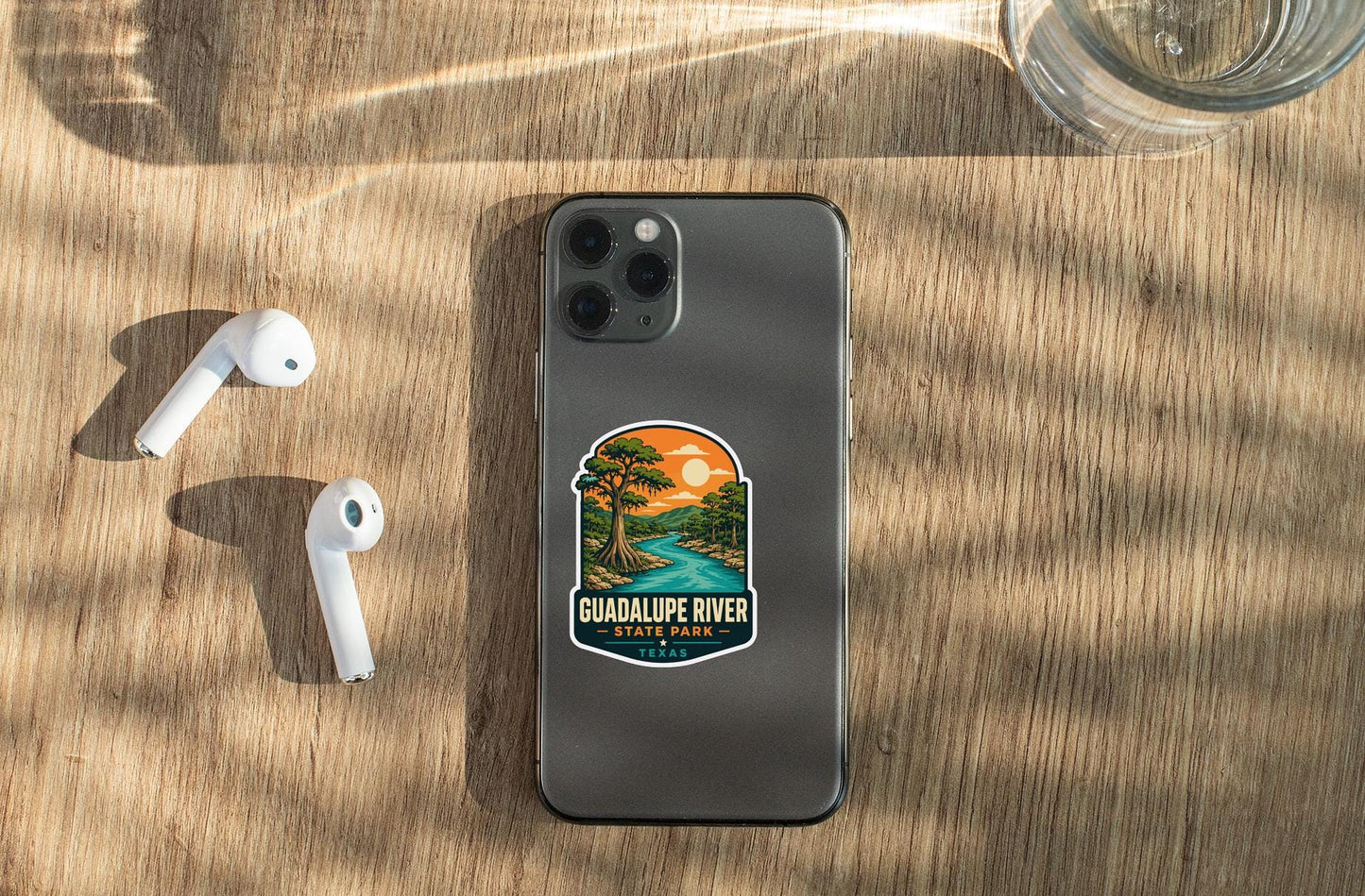 a smartphone with a sticker on the back, placed on a wooden surface alongside a pair of white earbuds and a glass of water.