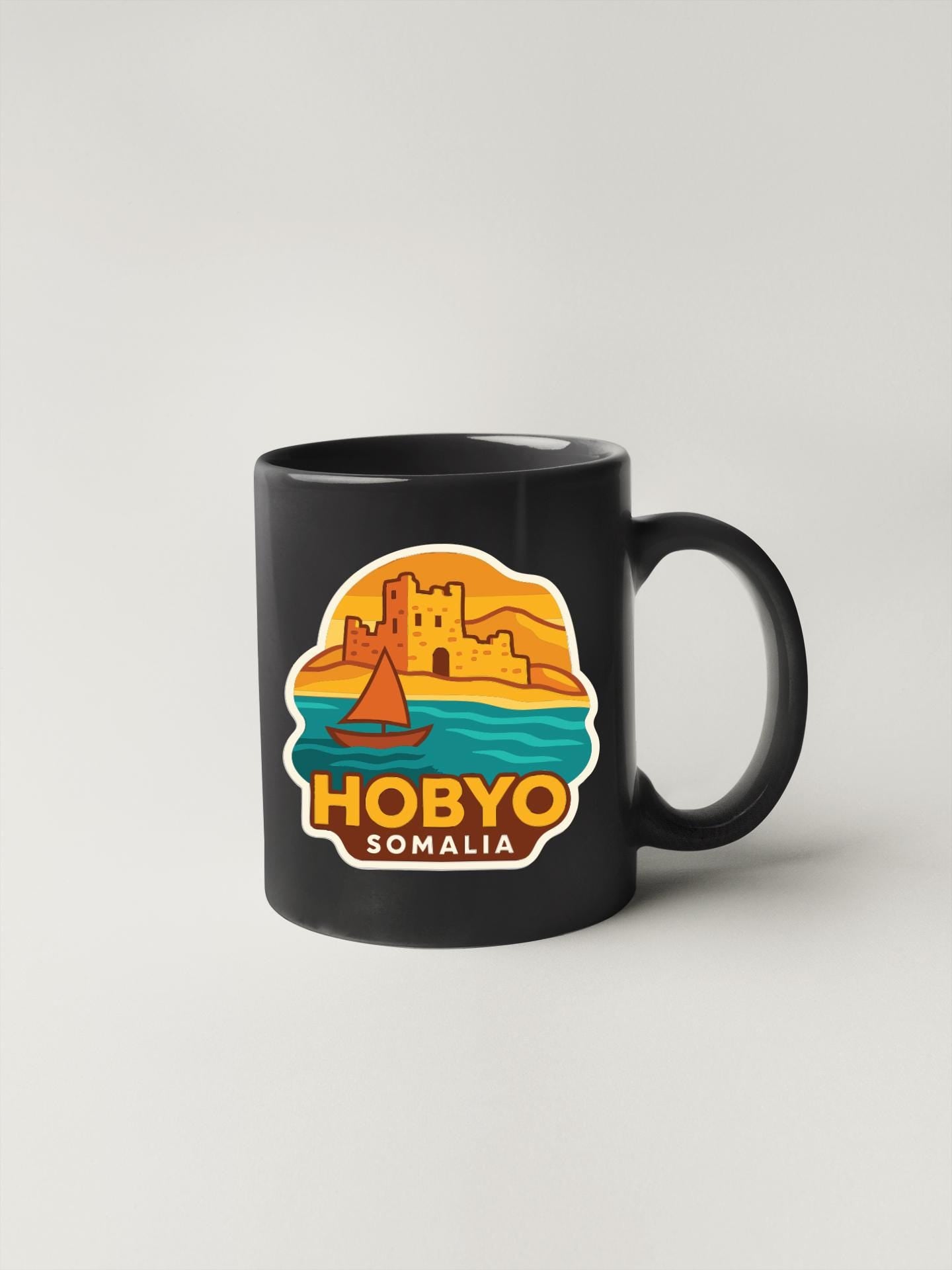 A black coffee mug with a colorful sticker depicting a castle and a sailboat on a body of water, along with the text "HOBBY SOMALIA".