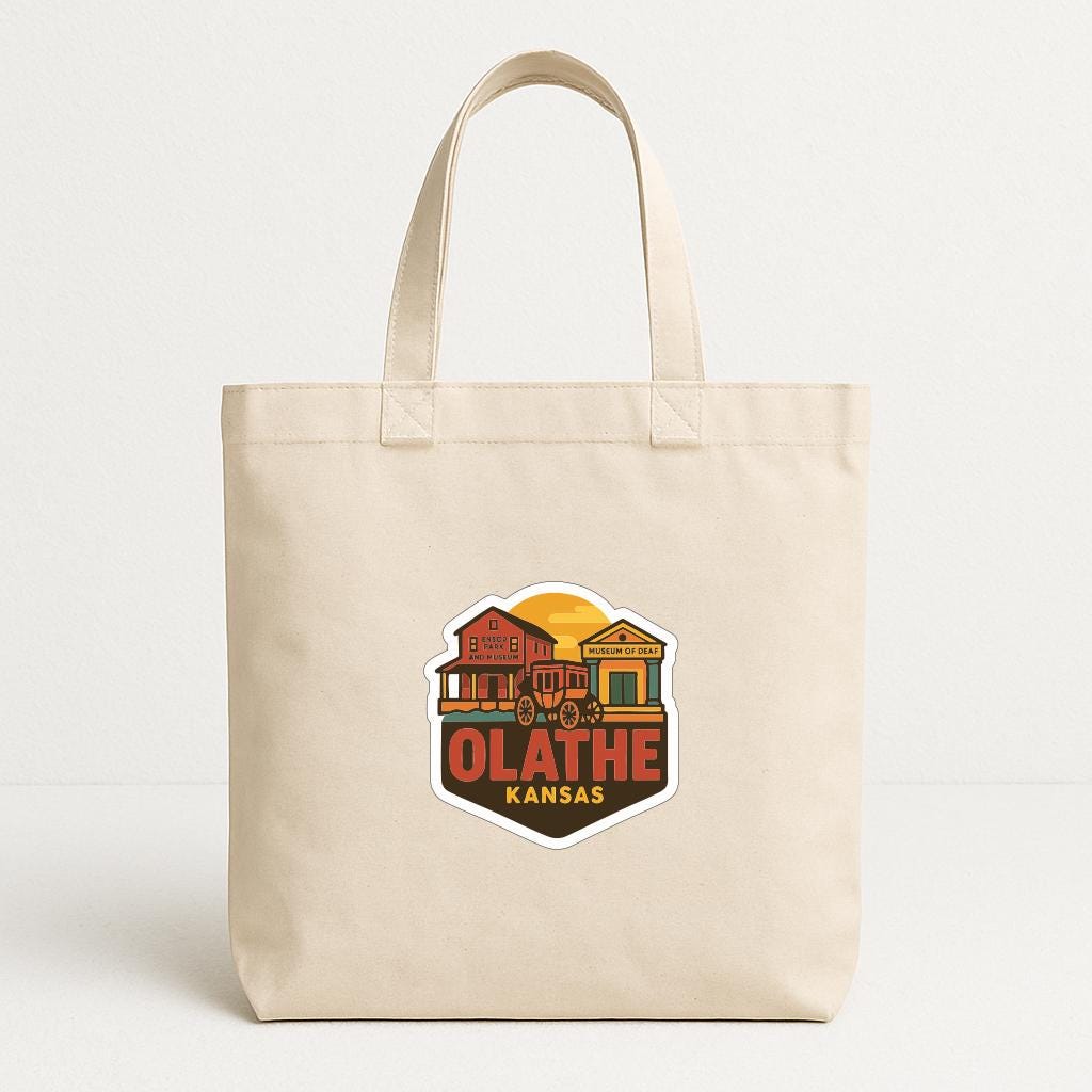 A beige tote bag with a graphic design featuring a stylized illustration of a town or city, along with the text "OLATHE KANSAS".