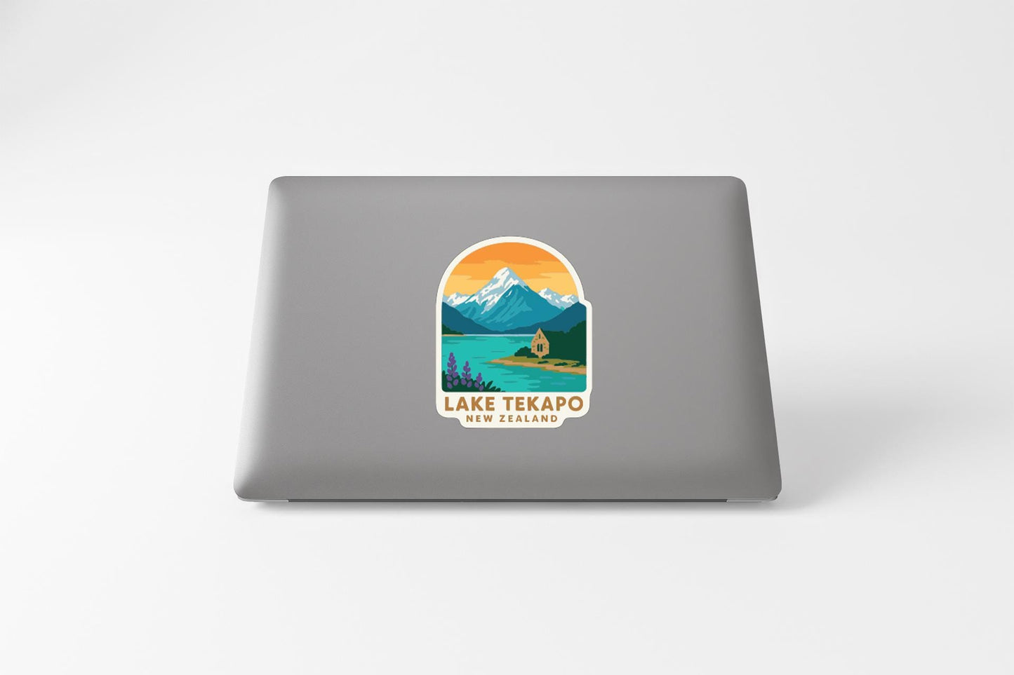 A sticker on the back of a laptop that features a mountain landscape with a lake and a cabin, along with the text "LAKE TEKAPO NEW ZEALAND".