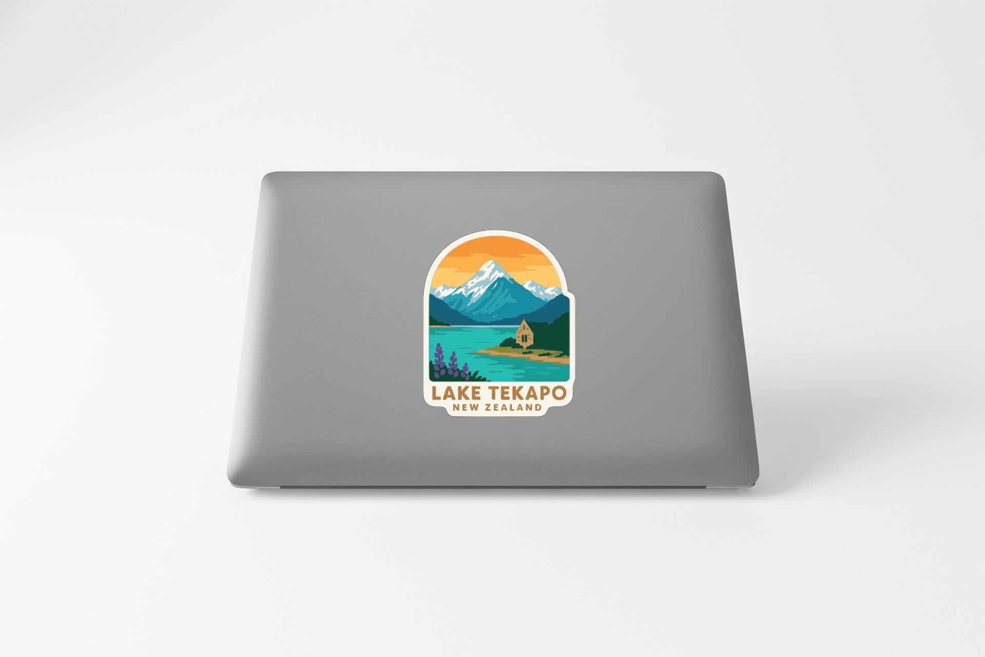 A sticker on the back of a laptop that features a mountain landscape with a lake and a cabin, along with the text "LAKE TEKAPO NEW ZEALAND".