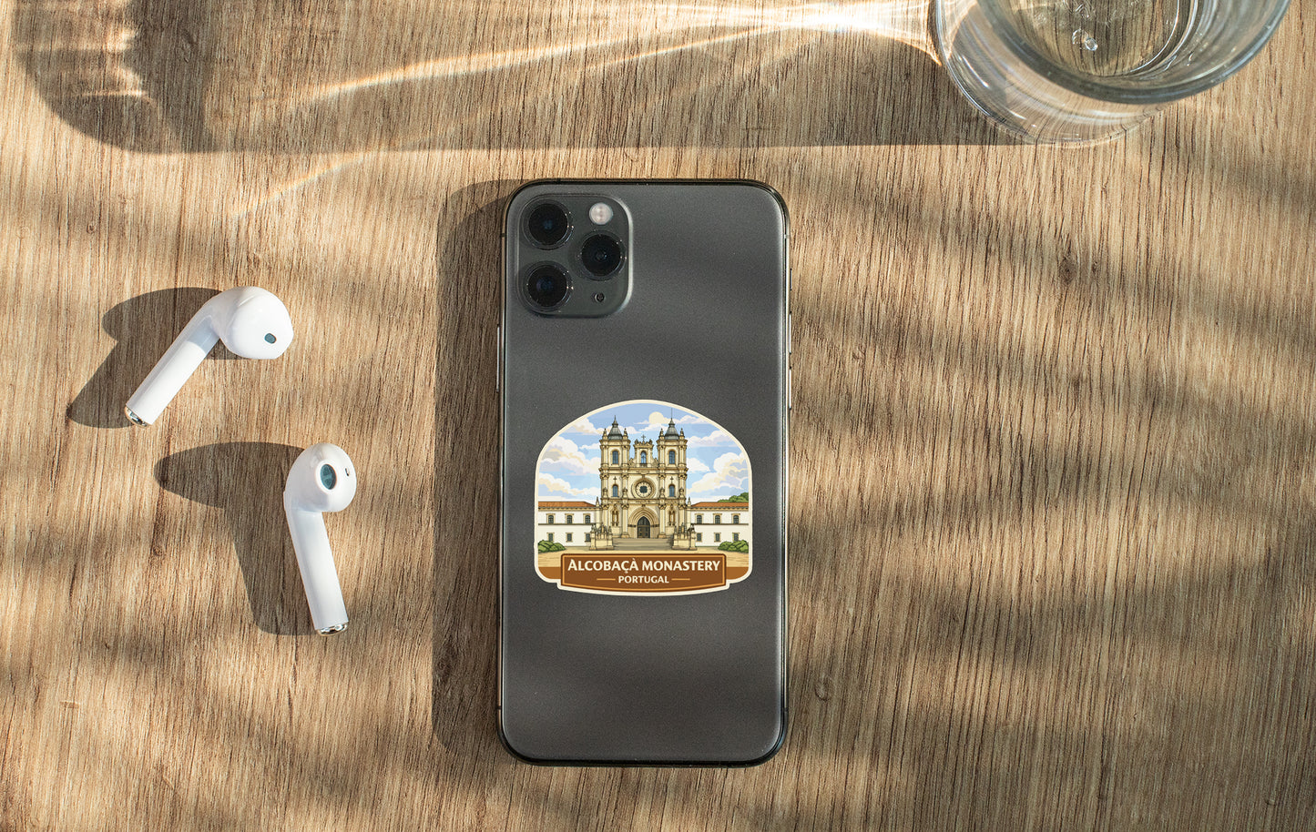 a smartphone with a sticker of a building on it, placed on a wooden surface alongside a pair of white earbuds and a glass of water.
