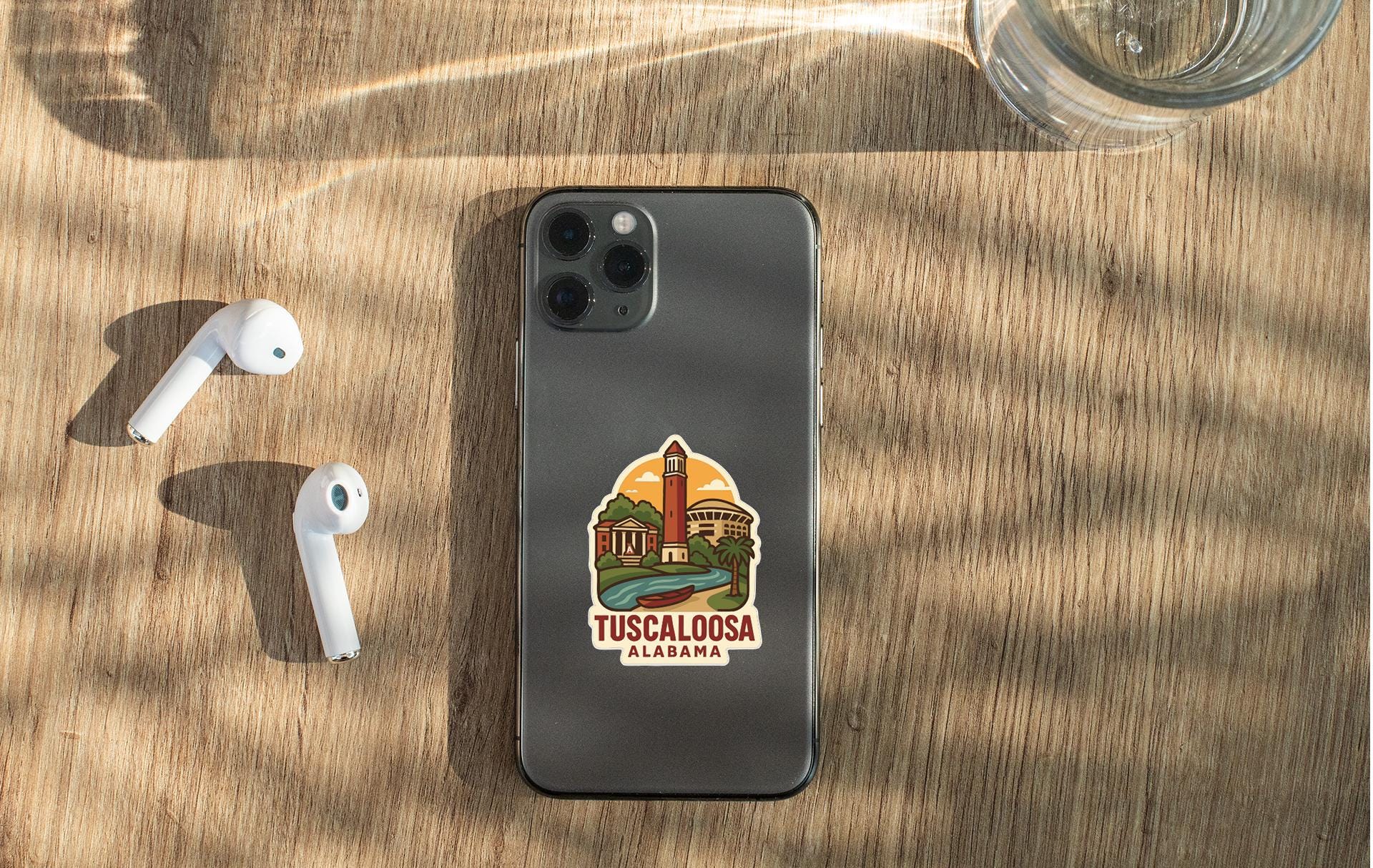 a smartphone with a sticker of Tuscalosa, Alabama, placed on a wooden surface alongside a pair of white earbuds and a glass of water.