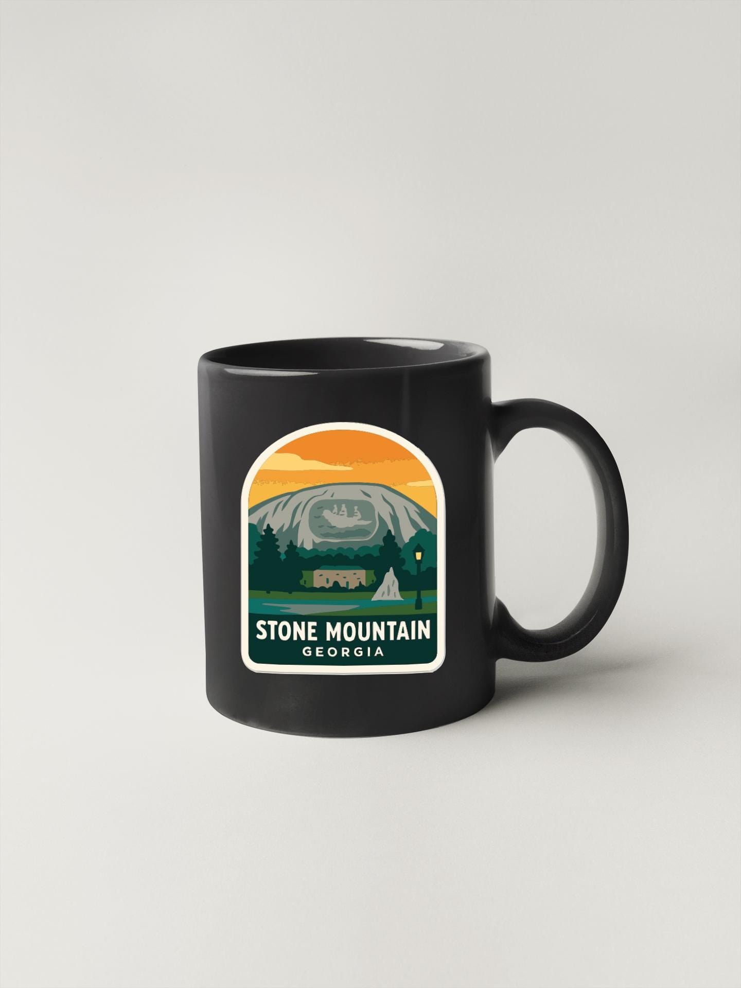 A black coffee mug with a sticker depicting a scenic mountain landscape in Stone Mountain, Georgia.