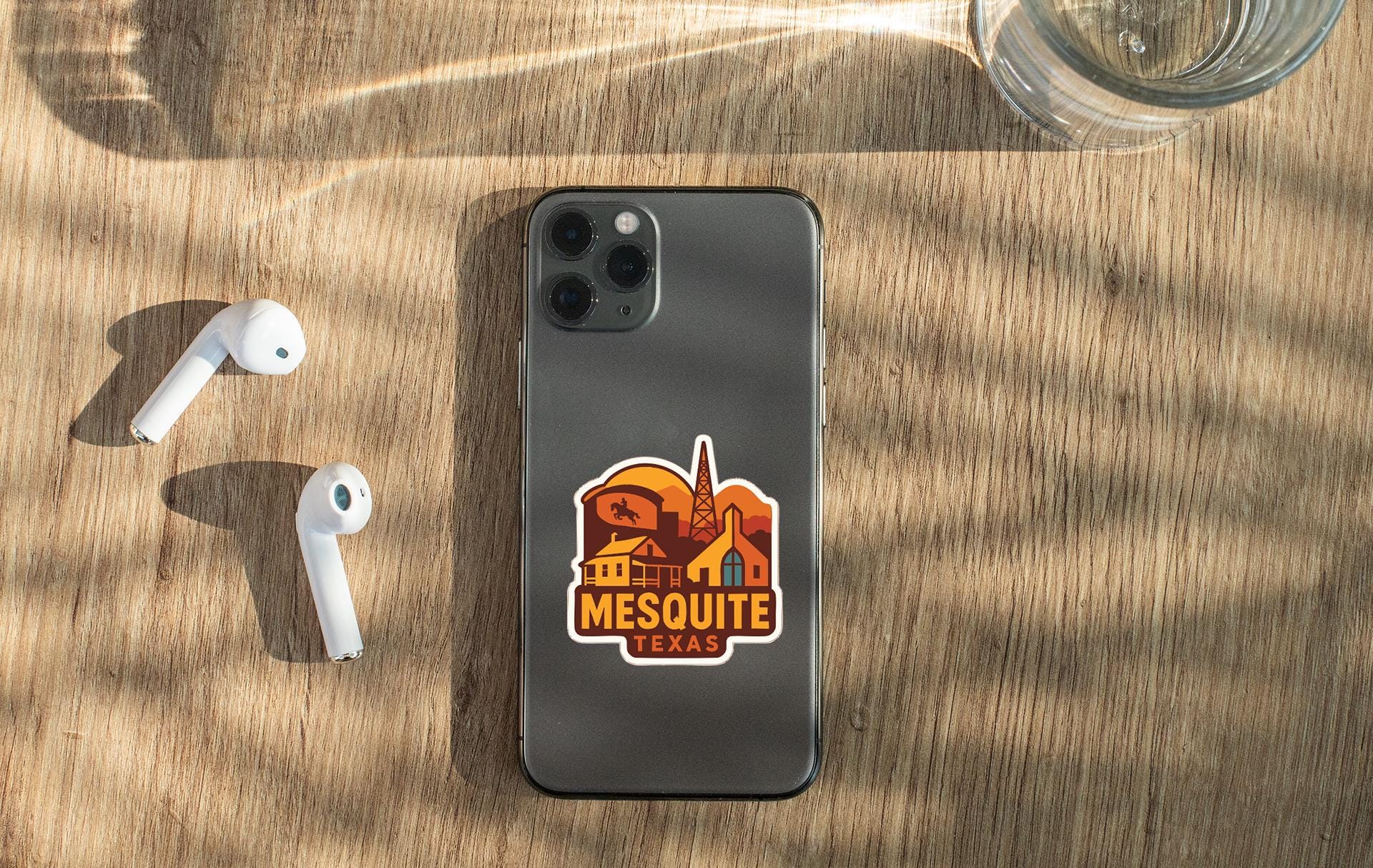 a smartphone with a sticker on the back, along with a pair of white earbuds and a glass of water on a wooden surface.