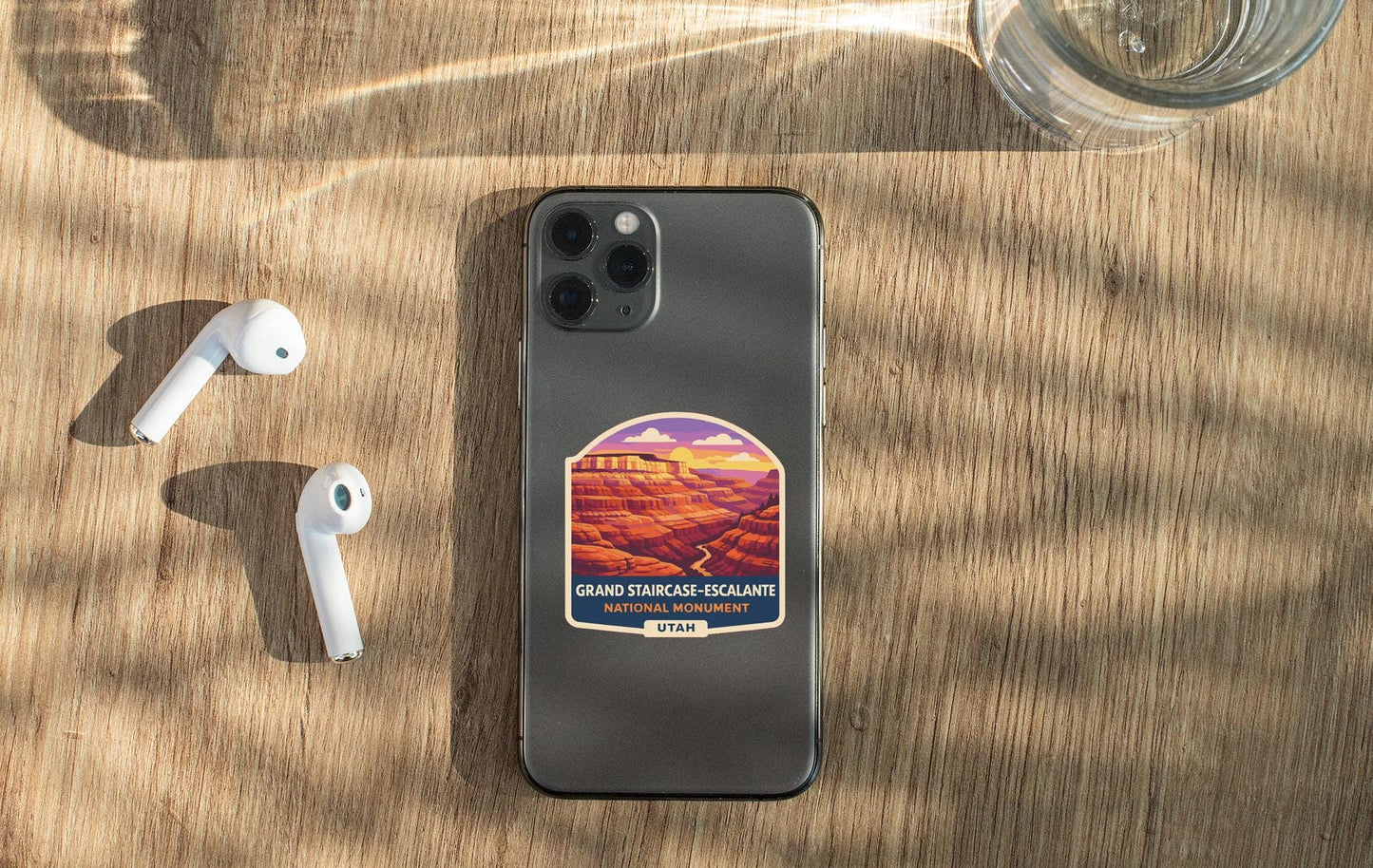 a smartphone with a sticker of a scenic mountain landscape, a pair of white earbuds, and a glass of water on a wooden surface.