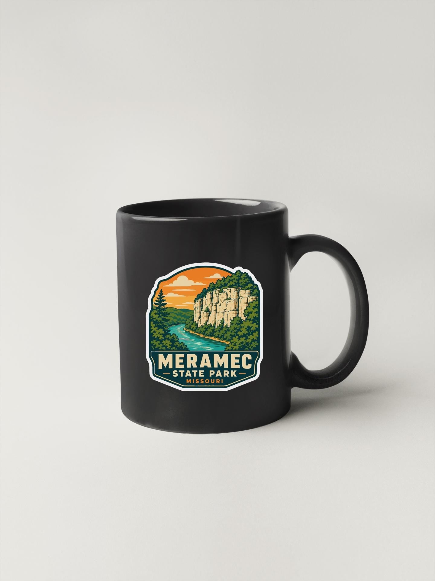 A black coffee mug with a graphic design depicting a scenic landscape, including mountains, a river, and a sunset.