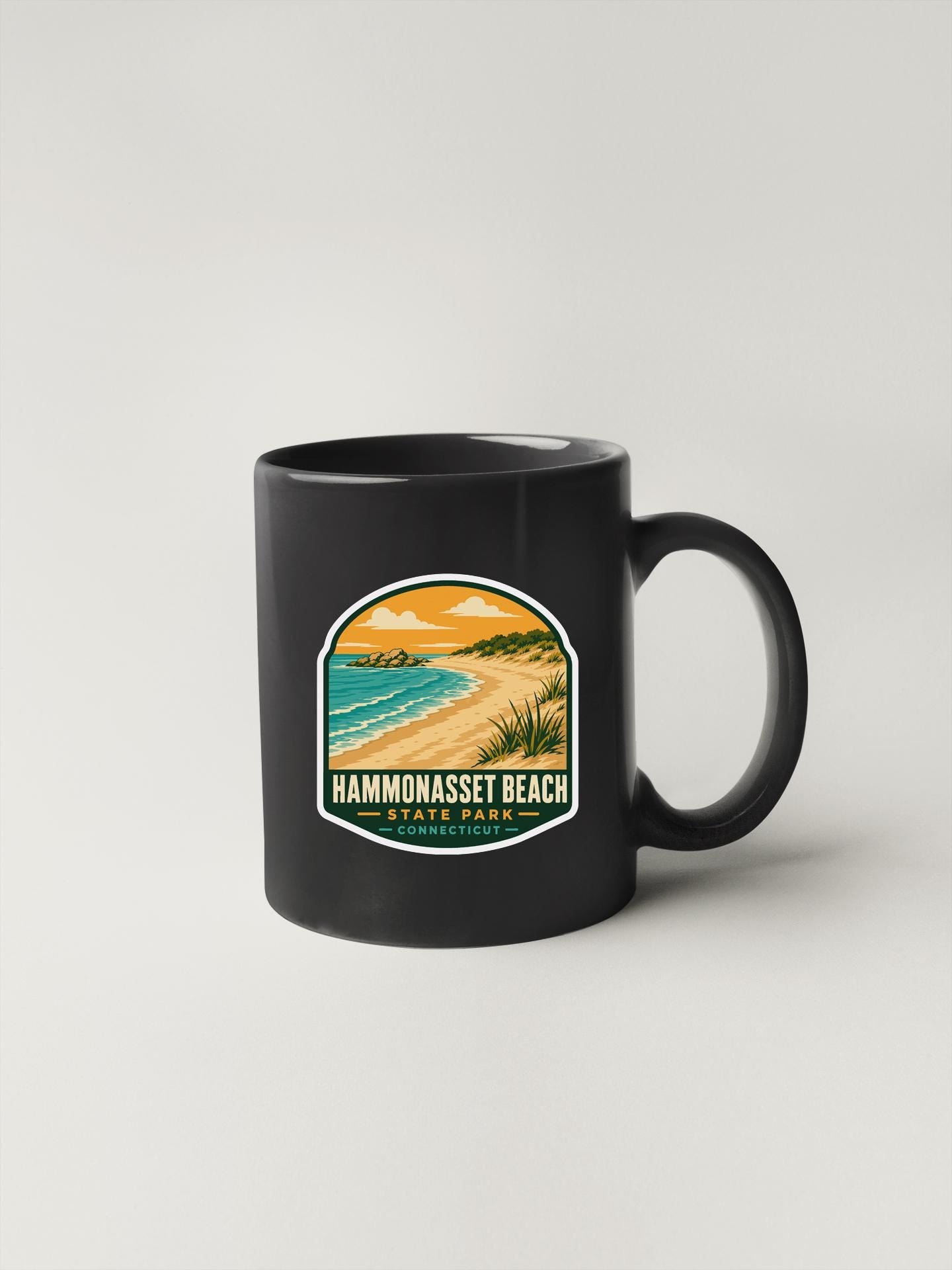A black coffee mug with a sticker depicting a beach scene and the text "HAMMONASSET BEACH STATE PARK" printed on it.