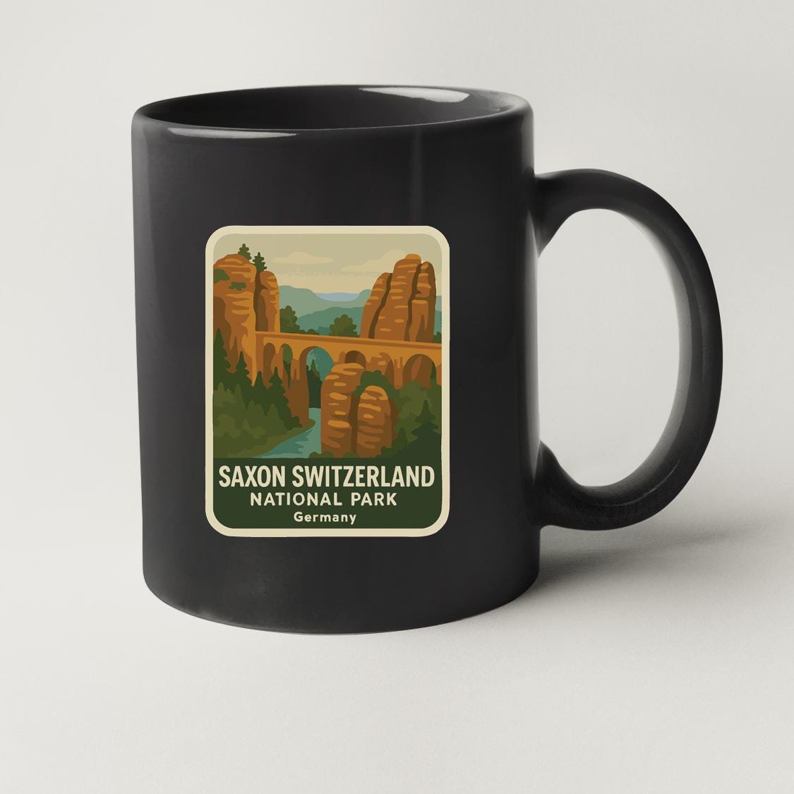 A black coffee mug with a sticker depicting the Saxon Switzerland National Park in Germany.