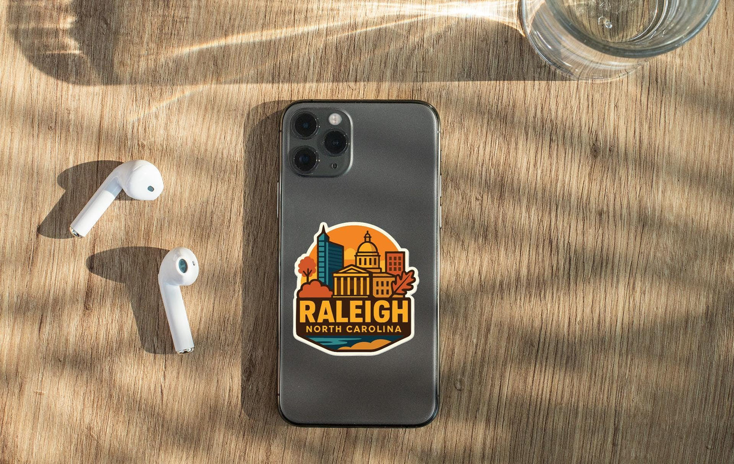 a smartphone with a sticker of Raleigh, North Carolina, on its back, along with a pair of white earbuds and a glass of water on a wooden surface.
