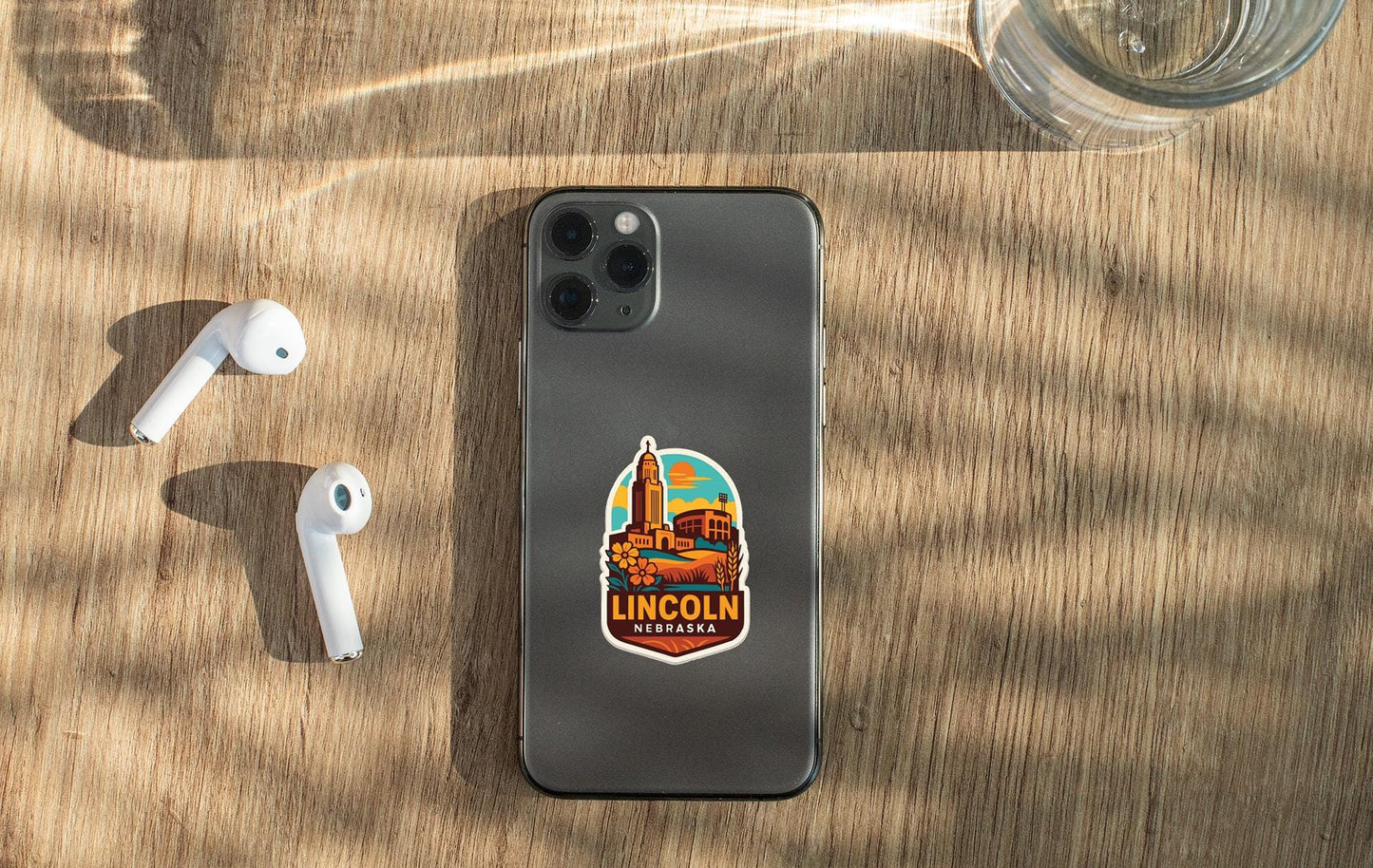 a smartphone with a sticker of Lincoln, Nebraska, on its back, along with a pair of white earbuds and a glass of water on a wooden surface.