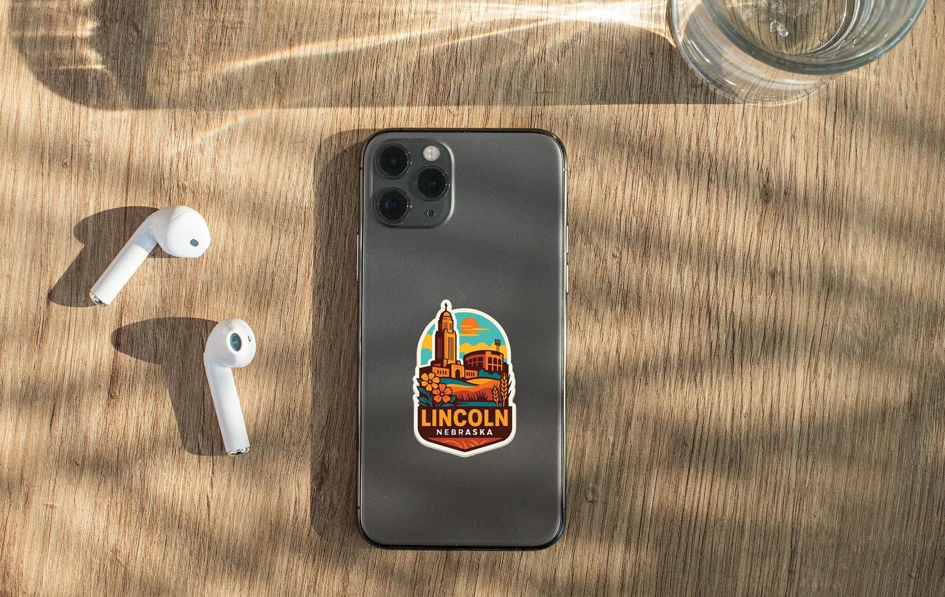 a smartphone with a sticker of Lincoln, Nebraska, on its back, along with a pair of white earbuds and a glass of water on a wooden surface.