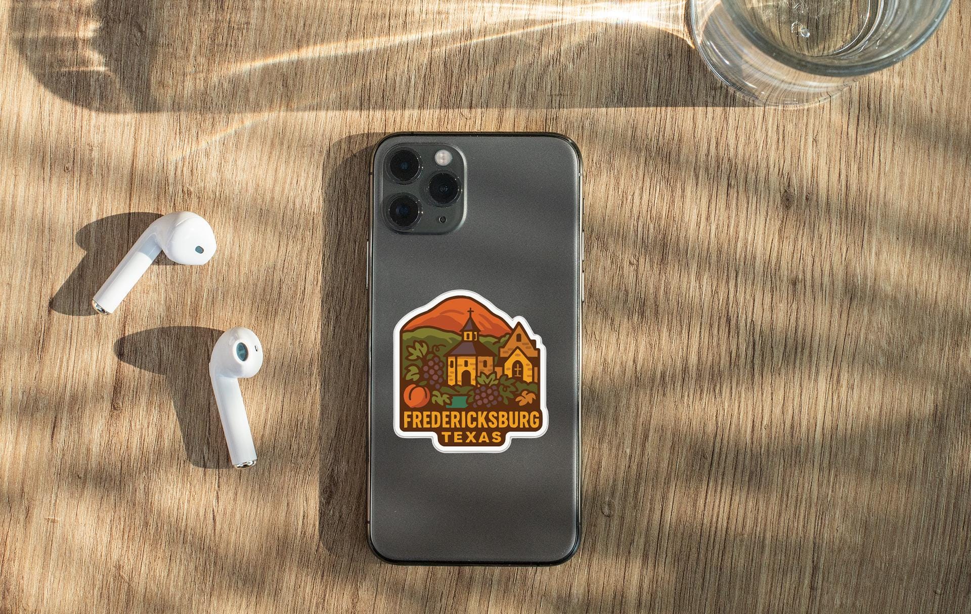 a smartphone with a sticker on the back, placed on a wooden surface alongside a pair of white earbuds and a glass of water.