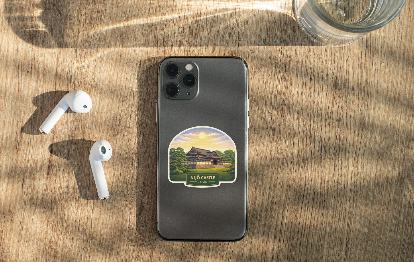 a smartphone with a sticker of a building, a pair of white earbuds, and a glass of water on a wooden surface.