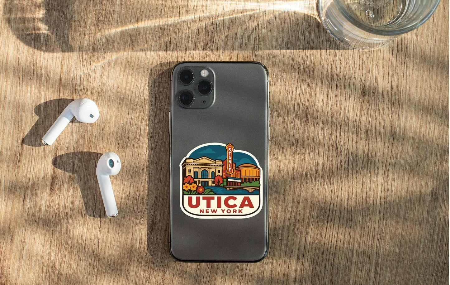 a smartphone with a sticker on the back, placed on a wooden surface alongside a pair of white earbuds and a glass of water.