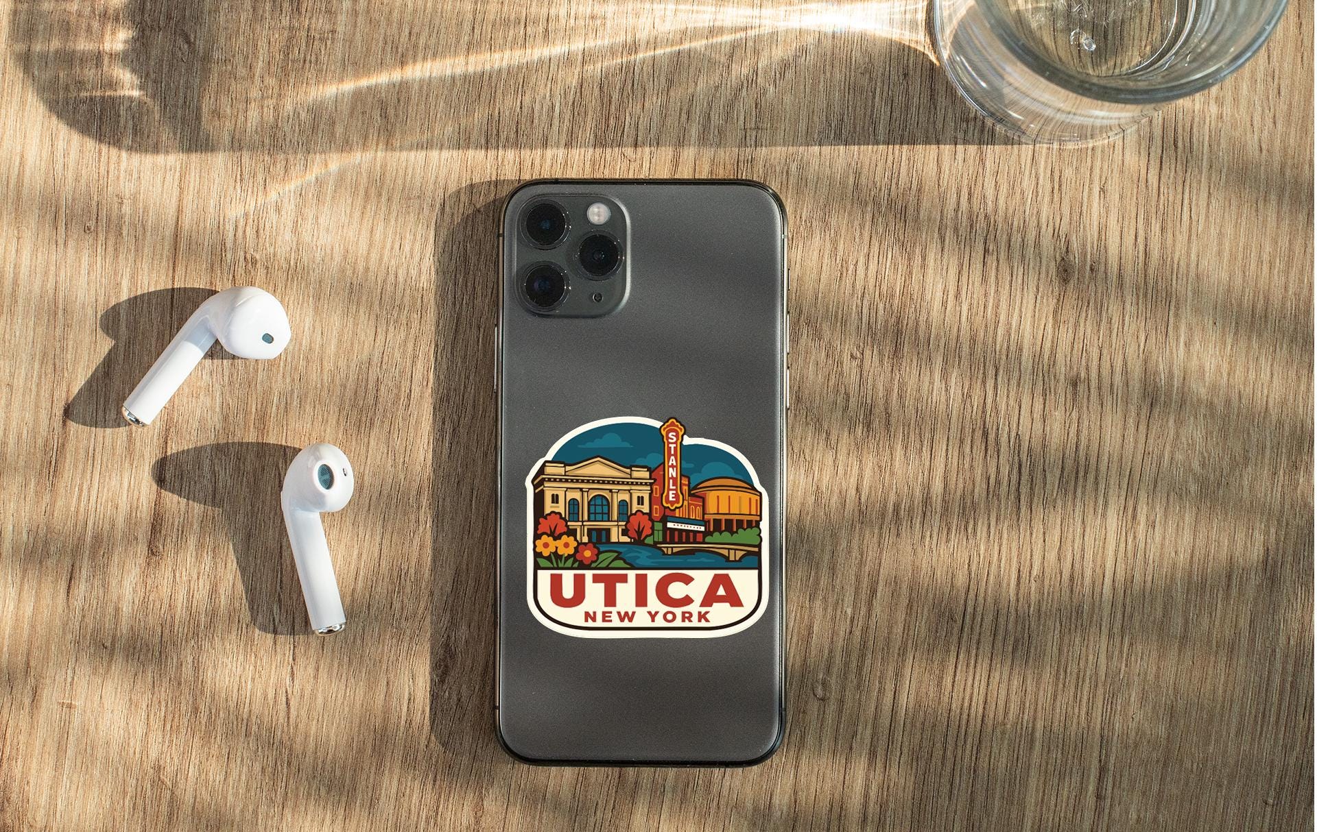 a smartphone with a sticker on the back, placed on a wooden surface alongside a pair of white earbuds and a glass of water.