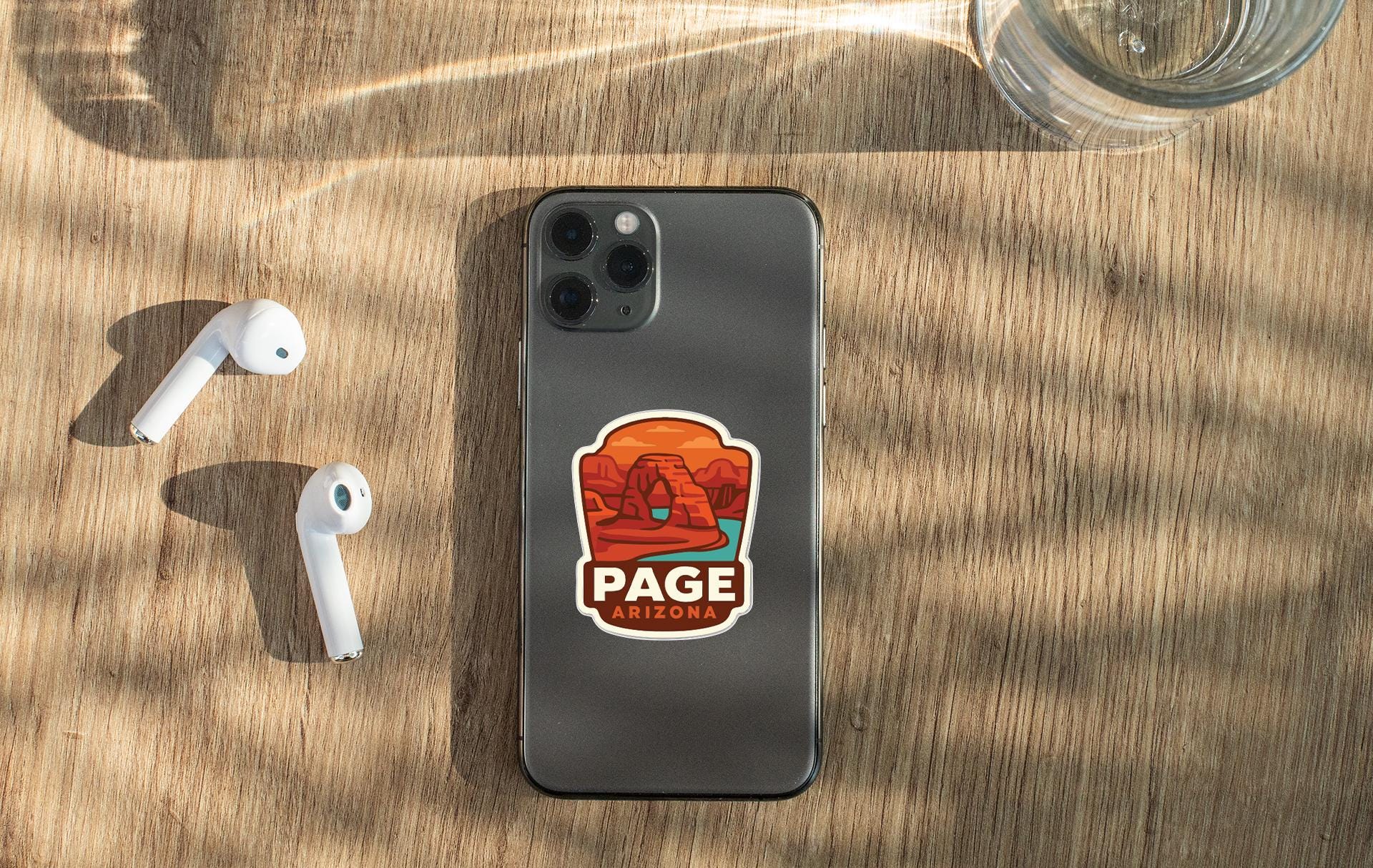a smartphone with a sticker of a desert landscape on it, along with a pair of white earbuds and a glass of water on a wooden surface.