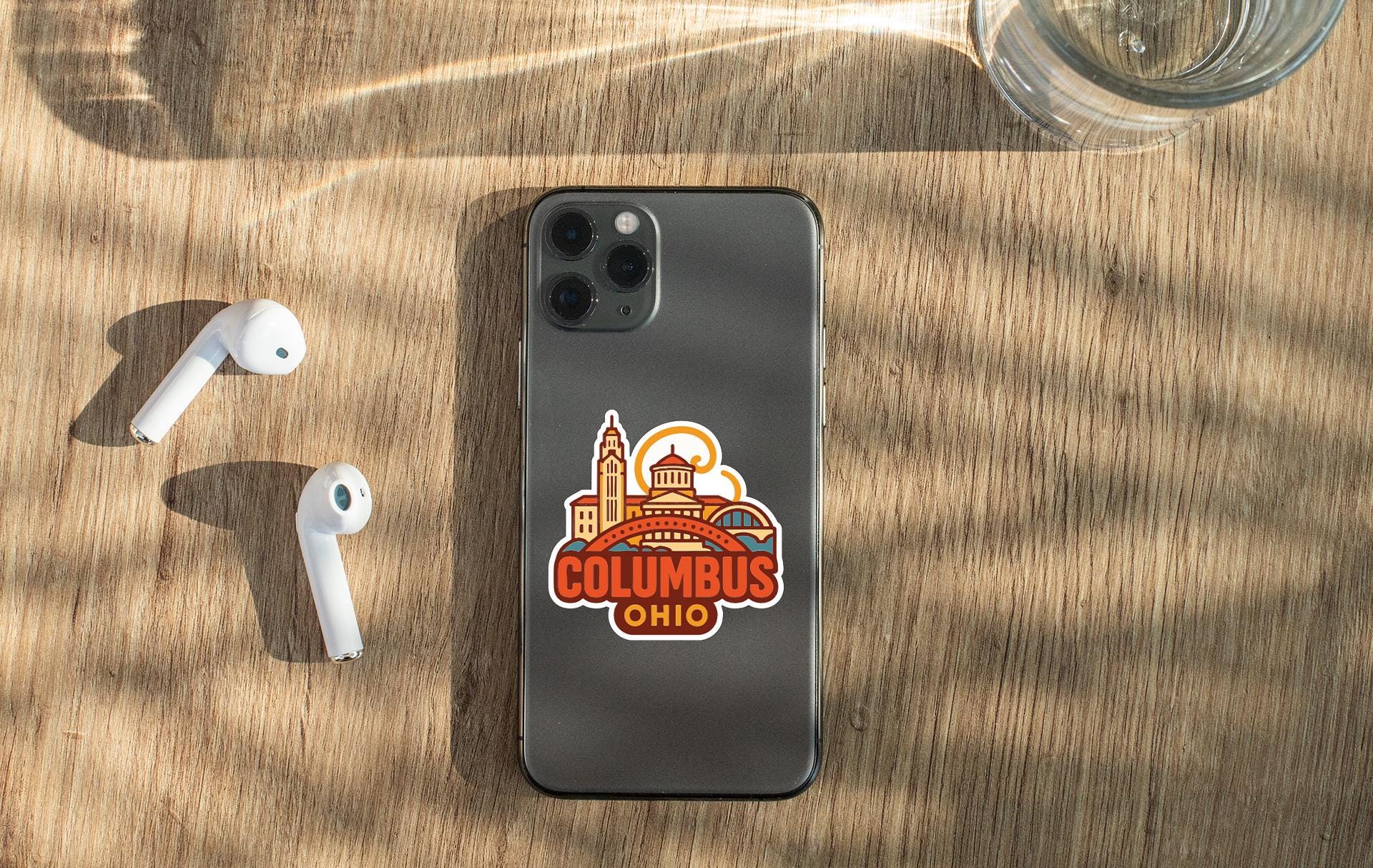 a smartphone with a sticker of Columbus, Ohio, placed on a wooden surface alongside a pair of white earbuds and a glass of water.