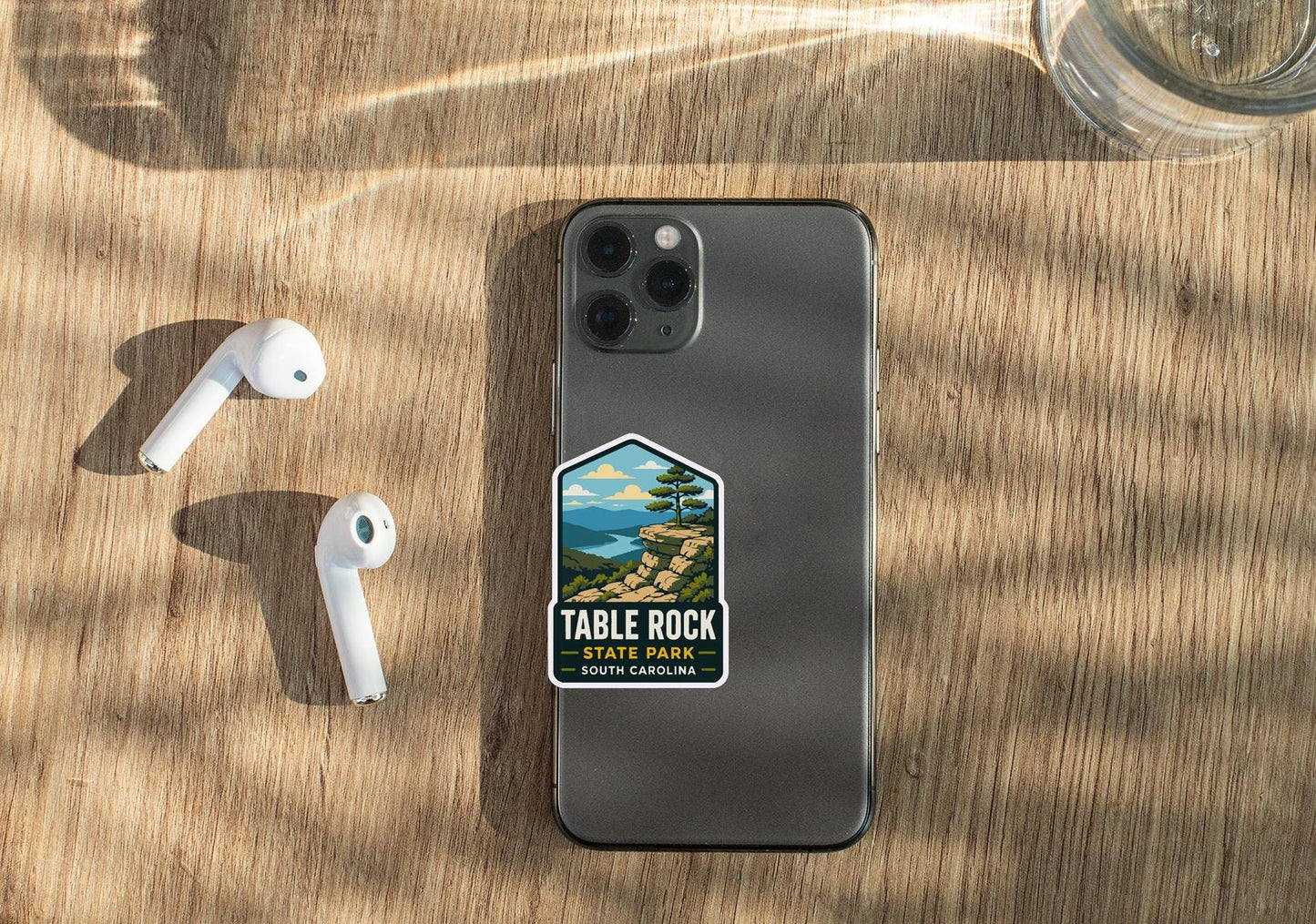 a smartphone with a sticker on the back, placed on a wooden surface alongside a pair of white earbuds and a glass of water.