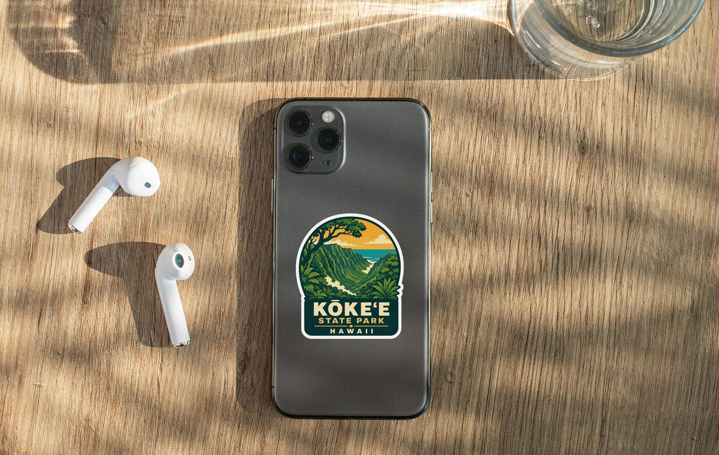 a smartphone with a sticker on the back, placed on a wooden surface alongside a pair of white earbuds and a glass of water.