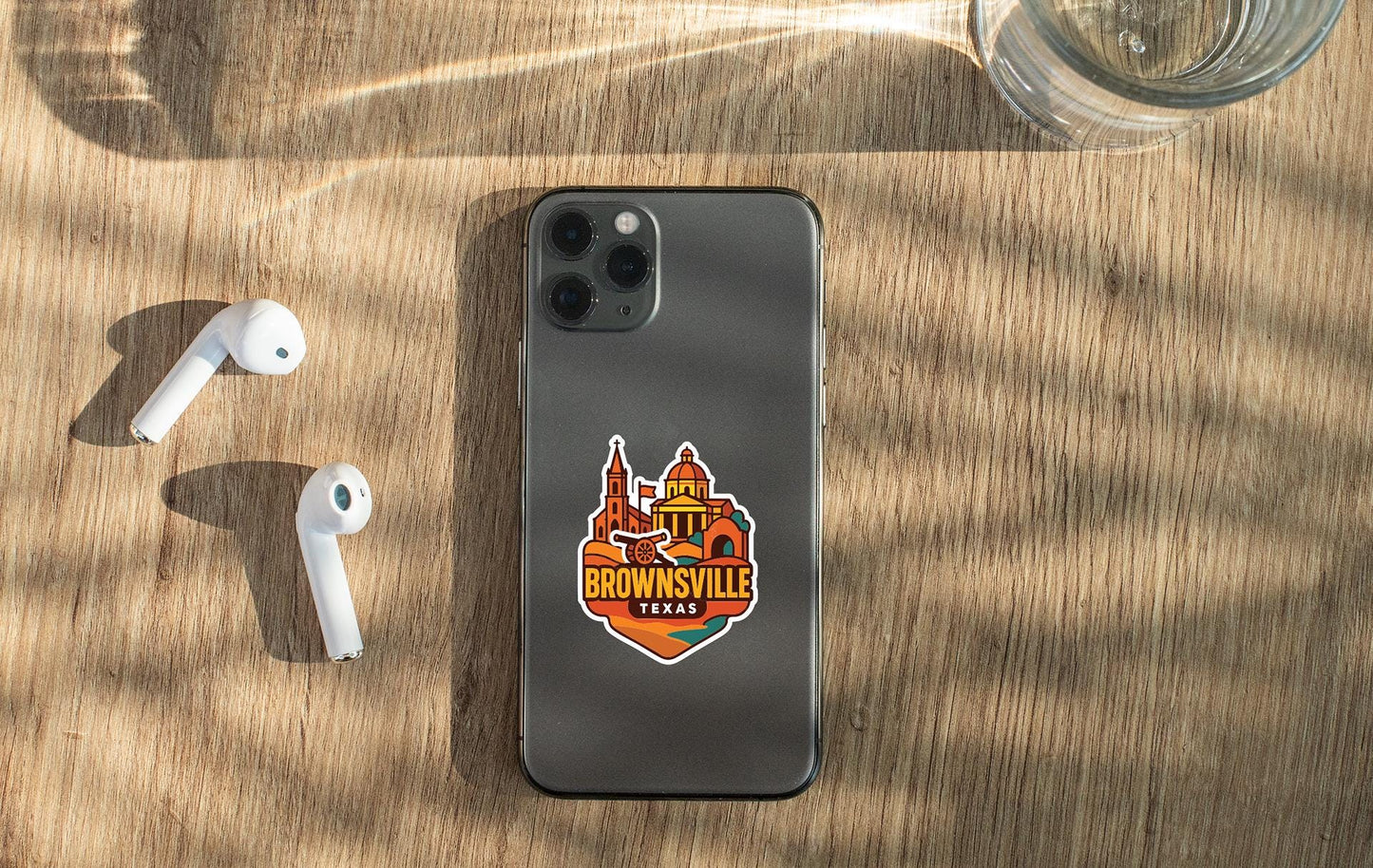 a smartphone with a sticker on the back, placed on a wooden surface alongside a pair of white earbuds and a glass of water.