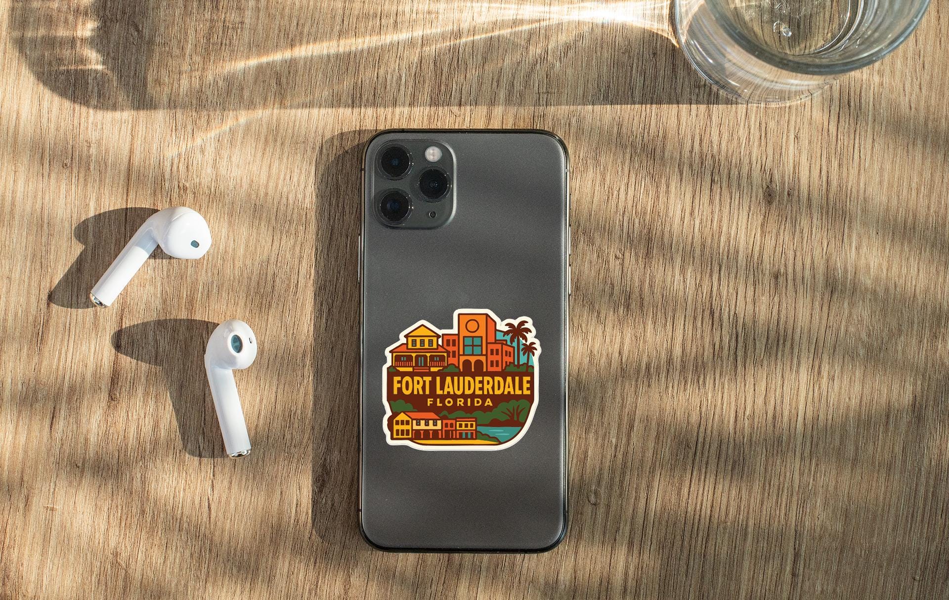 a smartphone with a sticker of Fort Lauderdale, Florida, placed on a wooden surface alongside a pair of white earbuds and a glass of water.