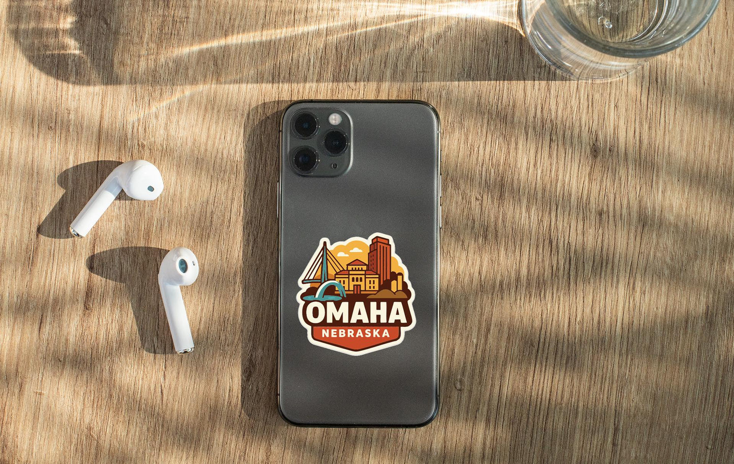 a smartphone with a sticker of Omaha, Nebraska, on its back, along with a pair of white earbuds and a glass of water on a wooden surface.