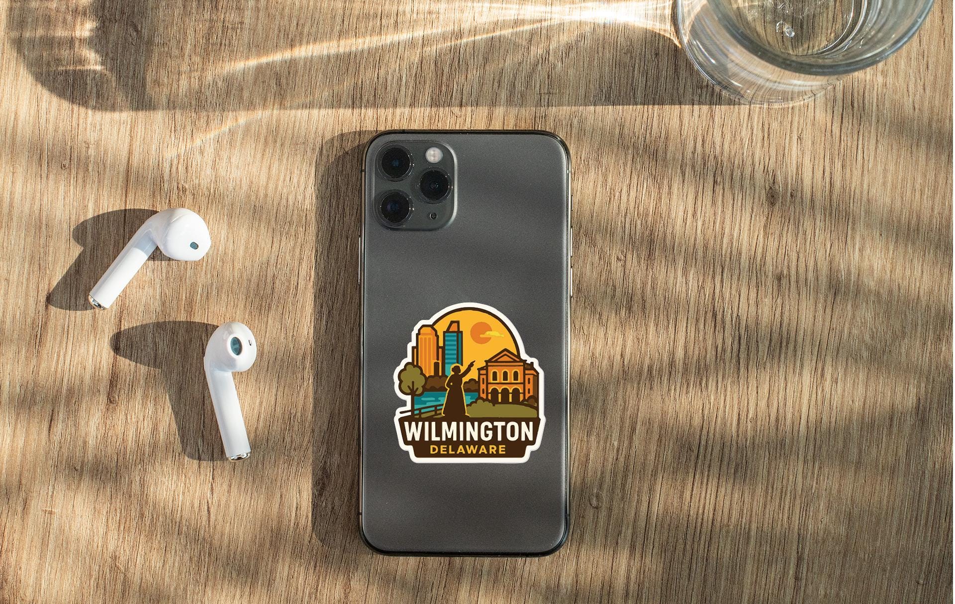 a smartphone with a sticker of Wilmington, Delaware, placed on a wooden surface alongside a pair of white earbuds and a glass of water.