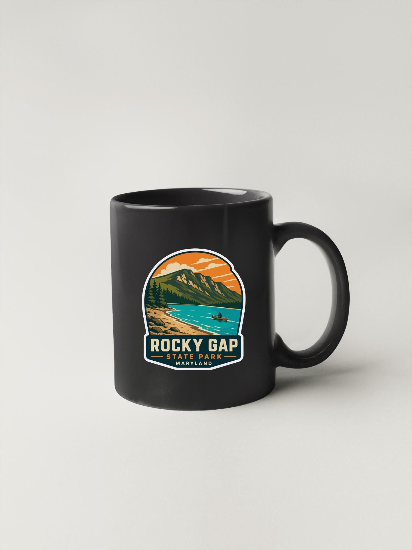A black coffee mug with a graphic design depicting Rocky Gap State Park in Maryland.