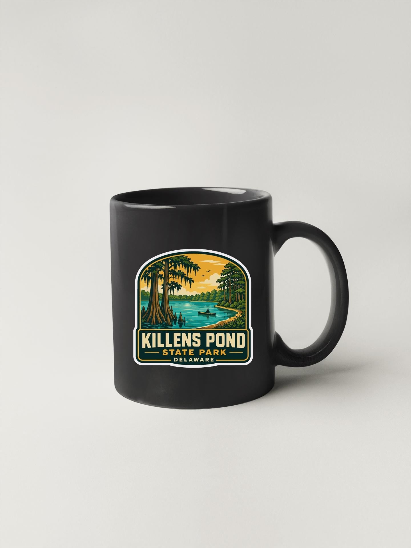 A black coffee mug with a graphic design depicting a scenic landscape featuring a pond, trees, and a sunset.