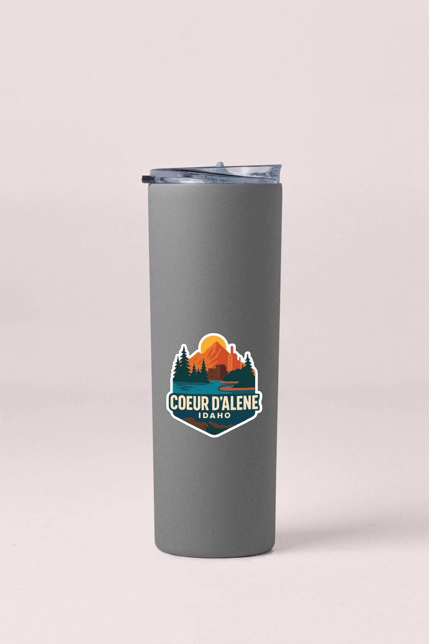 A gray insulated tumbler with a lid and a sticker featuring a mountain landscape and the text "COEUR D'ALENE IDAO".
