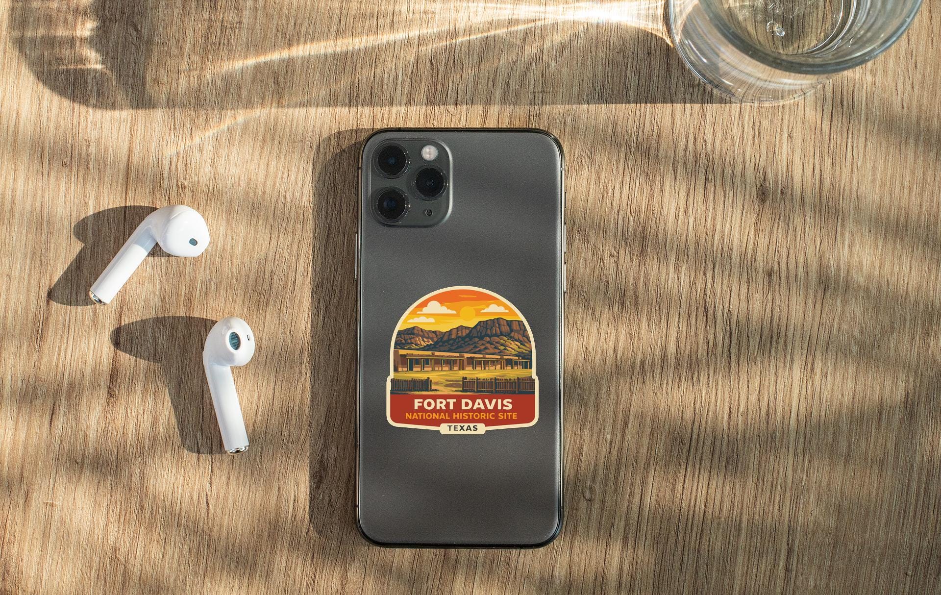 a smartphone with a sticker on the back, placed on a wooden surface alongside a pair of white earbuds and a glass of water.
