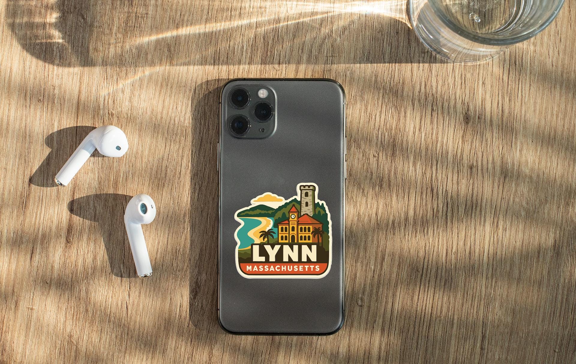 a smartphone with a sticker of Lynn, Massachusetts, placed on a wooden surface alongside a pair of white earbuds and a glass of water.
