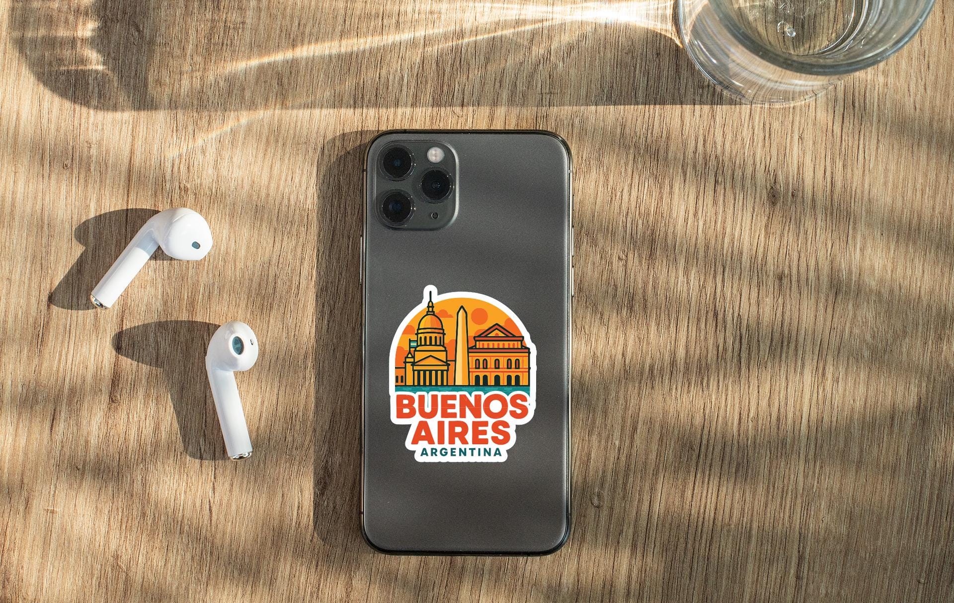 a smartphone with a sticker on the back, placed on a wooden surface alongside a pair of white earbuds and a glass of water.
