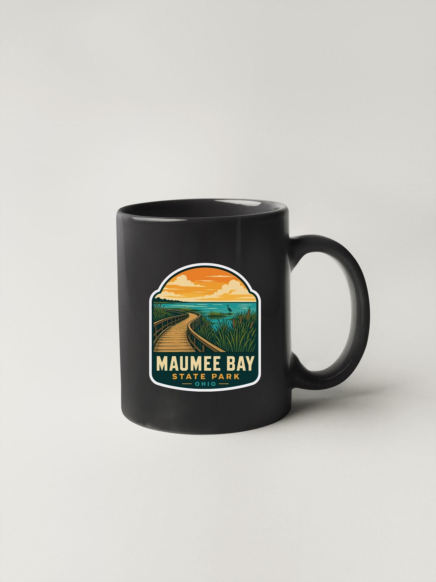 A black coffee mug with a graphic design depicting a scenic view of Maunee Bay State Park, featuring a winding path and a sunset landscape.