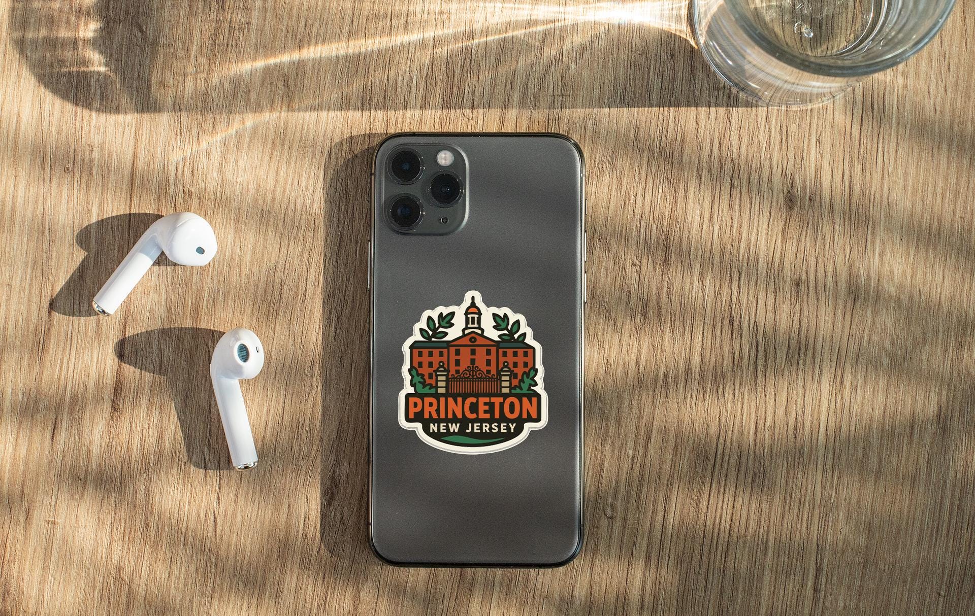 a smartphone with a Princeton New Jersey sticker on it, placed on a wooden surface alongside a pair of white earbuds and a glass of water.