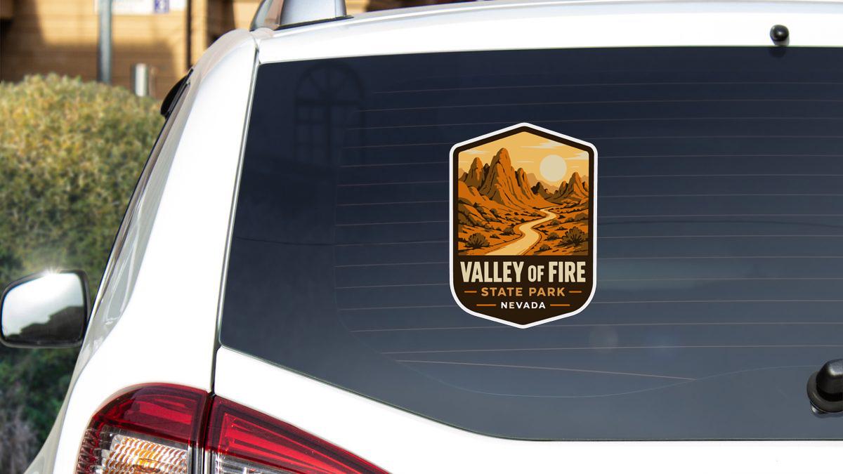 A sticker on the rear window of a white car, depicting a scenic landscape with mountains and a winding road, indicating the location of Valley of Fire State Park in Nevada.